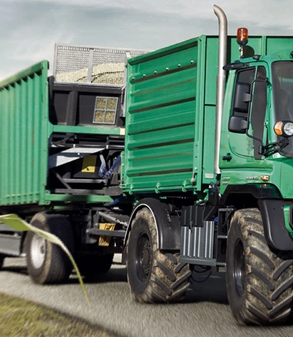 Grüner Unimog mit Anhänger auf ländlicher Straße.