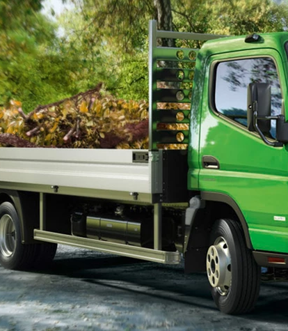Ein grüner Lastwagen transportiert Laub durch einen Park, während zwei Arbeiter mit Gartenarbeiten beschäftigt sind.
