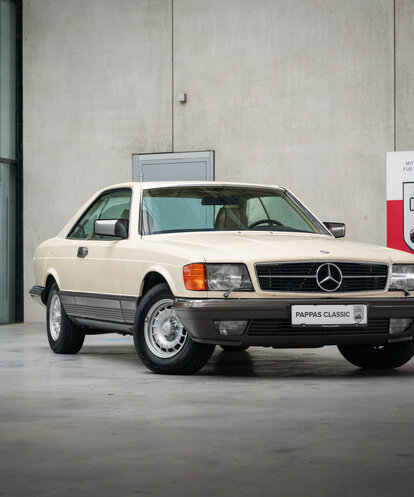 Ein cremefarbener Mercedes-Benz Oldtimer in einer modernen Ausstellungshalle mit dem Schild 'Pappas Classic'.