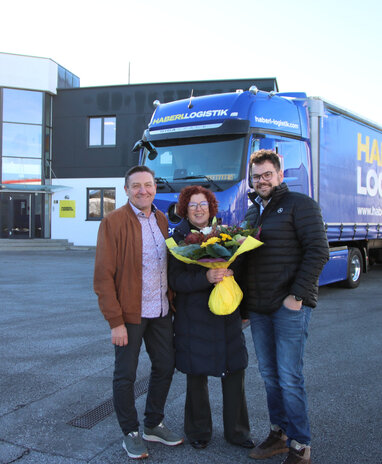 Zwei Männer und eine Frau mit Blumenstrauß vor einem blauen Haberl Logistik-Lkw.