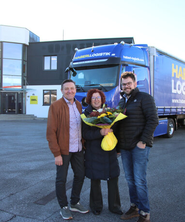 Zwei Männer und eine Frau mit Blumenstrauß vor einem blauen Haberl Logistik-Lkw.