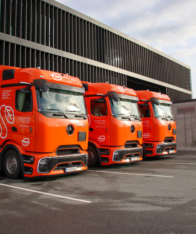 Drei orange Mercedes-Benz eActros, die an Ladestationen parken. | © Daimler Truck Austria