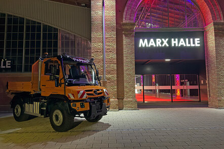Ein orangefarbener Mercedes-Benz Unimog steht vor der beleuchteten Marx Halle in einer nächtlichen Szene.