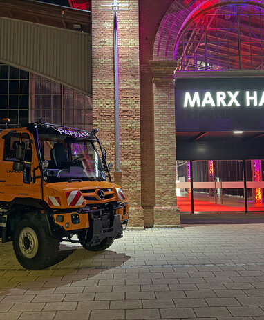Ein orangefarbener Mercedes-Benz Unimog steht vor der beleuchteten Marx Halle in einer nächtlichen Szene.