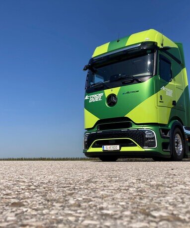 Ein grüner Mercedes-Benz eActros auf einer asphaltierten Fläche unter blauem Himmel.