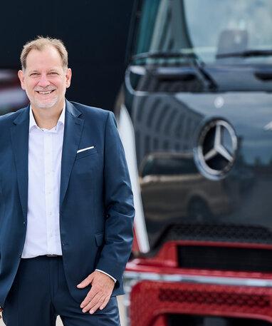 Bernhard Rusch steht im Anzug lächelnd vor einem Mercedes-Benz Lkw.