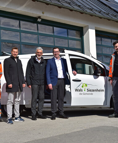 Personen vor einem Fahrzeug der Gemeinde Wals-Siezenheim, einer trägt eine orange Arbeitskleidung.
