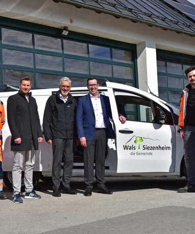 Personen vor einem Fahrzeug der Gemeinde Wals-Siezenheim, einer trägt eine orange Arbeitskleidung.