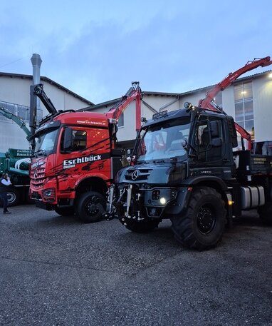 Ein roter Lkw und ein schwarzer Unimog mit Kranaufbauten stehen in der Abenddämmerung nebeneinander.