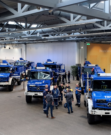 Mercedes-Benz Einsatzfahrzeuge bei einer Veranstaltung in einer großen Halle. Personen stehen um die Fahrzeuge herum. | © THW