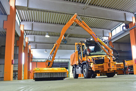 Oranges Straßenreinigungsfahrzeug in einer Garage, mit ausgefahrenem Mäharm und Wasserbehälter.