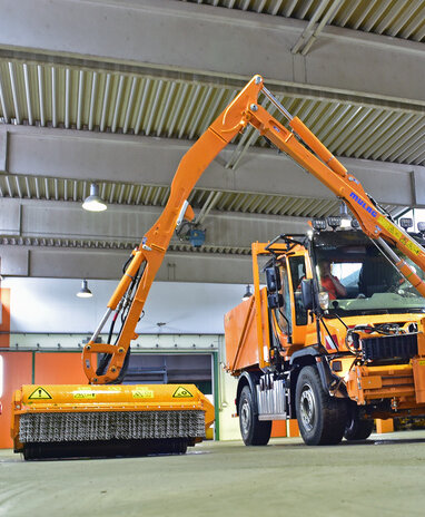Oranges Straßenreinigungsfahrzeug in einer Garage, mit ausgefahrenem Mäharm und Wasserbehälter.
