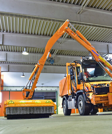 Oranges Straßenreinigungsfahrzeug in einer Garage, mit ausgefahrenem Mäharm und Wasserbehälter.