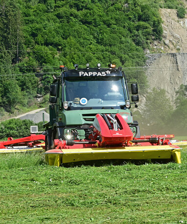 Ein Unimog mäht eine Wiese in einer bergigen Landschaft.