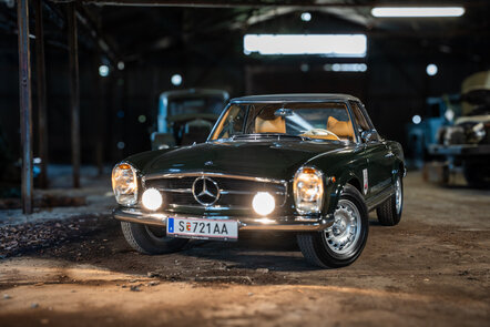 Ein dunkler, klassischer Mercedes-Benz 250 SL mit eingeschalteten Scheinwerfern in einer Werkstatt.