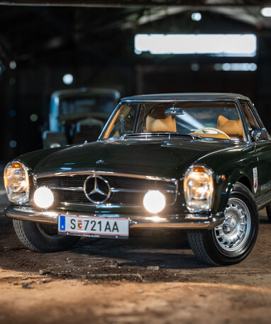 Ein dunkler, klassischer Mercedes-Benz 250 SL mit eingeschalteten Scheinwerfern in einer Werkstatt.