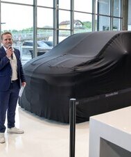 Ein Mann mit Mikrofon steht vor dem verhüllten Mercedes-Benz VLE und spricht zu den Gästen.