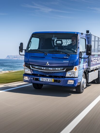 Blauer FUSO eCanter Lkw fährt auf einer Küstenstraße am Meer entlang.