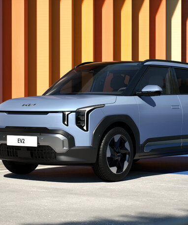 Silberner Kia EV2 SUV bei Sonnenschein vor bunter Streifenwand.