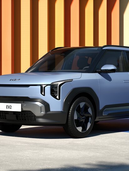 Silberner Kia EV2 SUV bei Sonnenschein vor bunter Streifenwand.