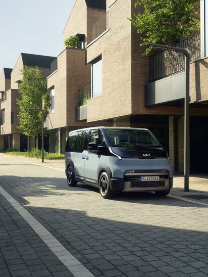 Blauer Kia PV5 Passenger in schräger Frontansicht zwischen modernen Wohnhäusern.