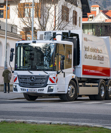Ein weißer Mercedes-Benz eEconic Müllwagen fährt durch eine städtische Umgebung. Aufschrift: "Voll elektrisch durch die Stadt".