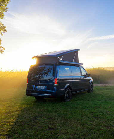 Ein blauer Mercedes-Benz Marco Polo mit Aufstelldach parkt auf einer Wiese am Ufer eines Sees im Morgenlicht. | © Mercedes-Benz AG