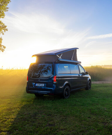 Ein blauer Mercedes-Benz Marco Polo mit Aufstelldach parkt auf einer Wiese am Ufer eines Sees im Morgenlicht. | © Mercedes-Benz AG