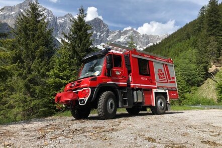 Rotes Feuerwehrfahrzeug in einer alpinen Gegend, umgeben von Bäumen und Bergen im Hintergrund.