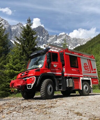 Rotes Feuerwehrfahrzeug in einer alpinen Gegend, umgeben von Bäumen und Bergen im Hintergrund.